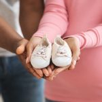 Parenthood concept. Closeup of cute baby shoes in pregnant black couple hands. Unrecognizable african american man and woman showing small shoes for their coming baby, cropped